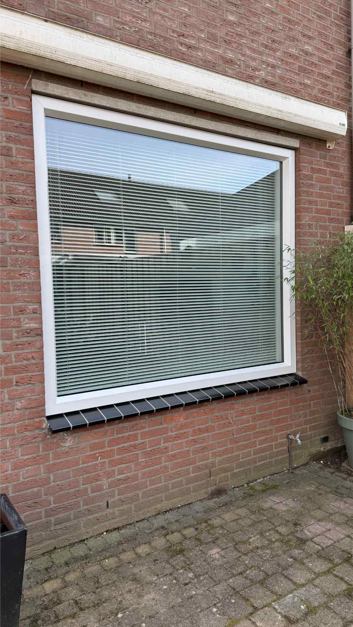 Wit kunststof kozijn in een bakstenen gevel met jaloezieën aan de binnenkant